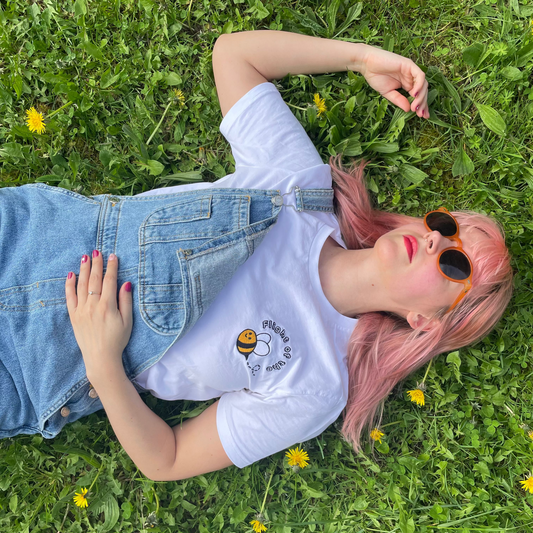 woman laying in grass with Jeans overalls and a white t-shirt from Cool Classical Studios with an embroidered Flight of the Bumblebee logo on the left chest. 
